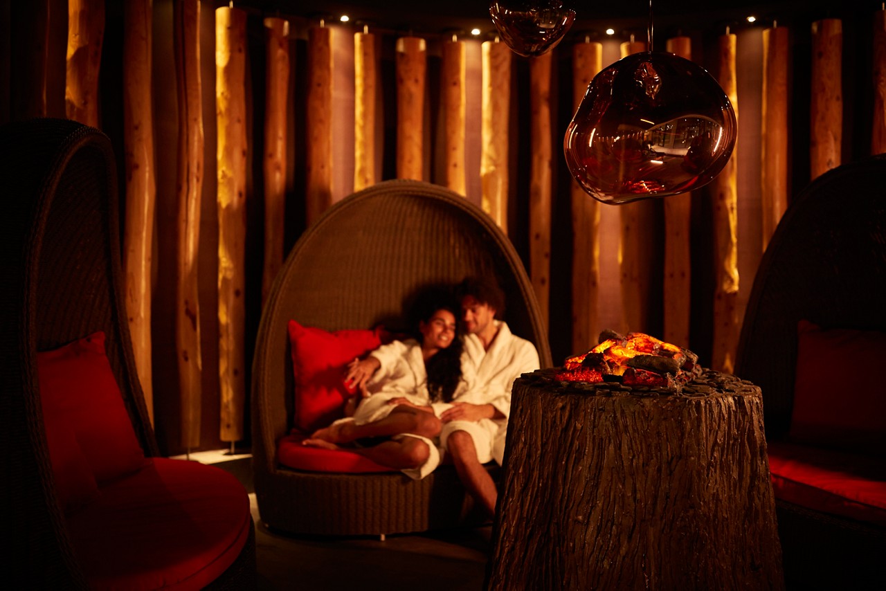 Man and woman snuggled up in a large cocoon chair by a glowing fire.